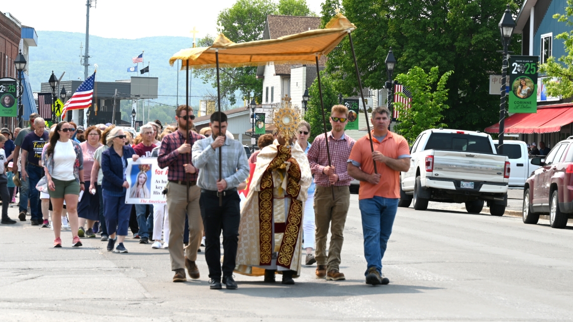 Father Antony Alexander Maria Doss carries a monstrance holding the Blessed Sacrament. Four men carry a canopy which he is walking beneath.