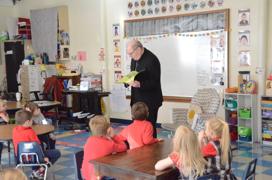 The bishop stands at the front of a classroom reading a book to seated children.