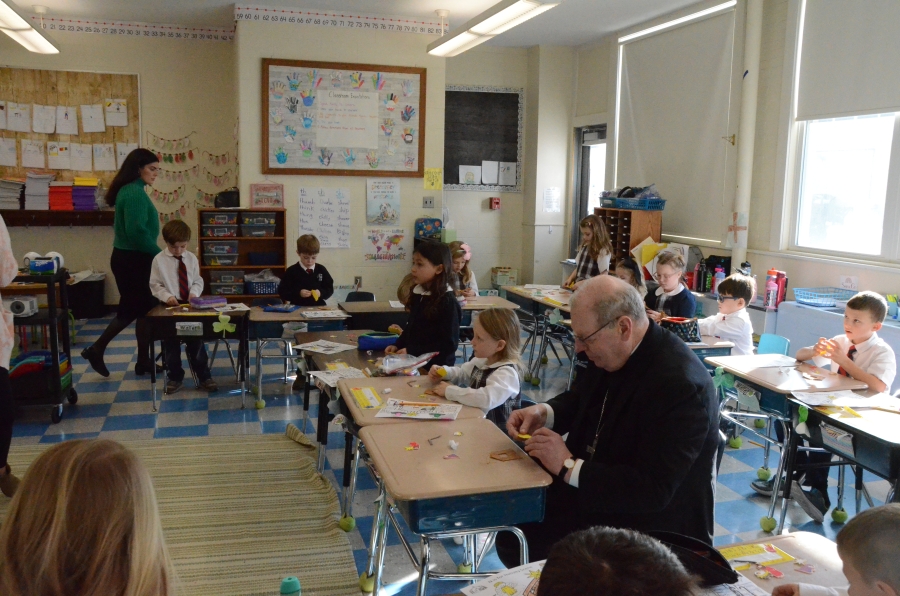 The bishop seated at a desk making an ornament with students.