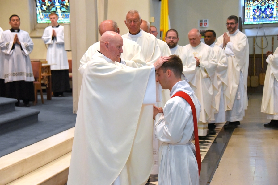 Father Joseph Ford lays hands on Deacon Matthew Valles with priests lined up in the background.