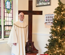 Father Seamus Griesbach standing next to a cross. A Christmas tree is in the corner.