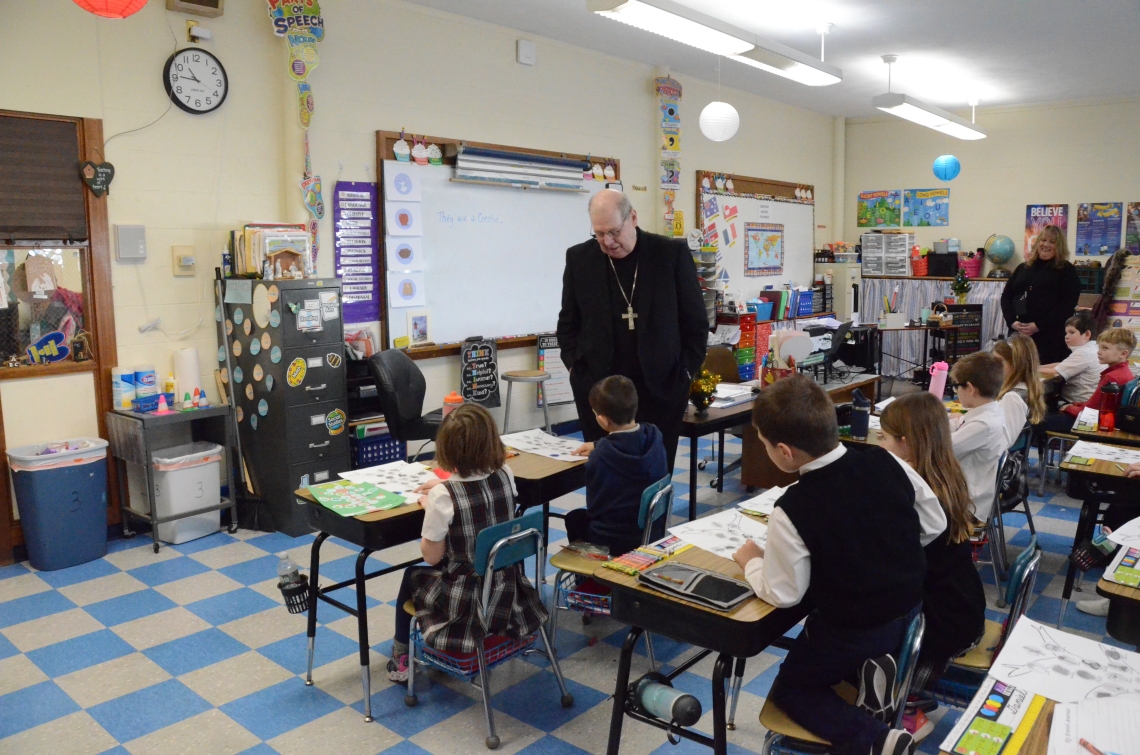 The bishop stands at the front of the classroom speaking to students sitting at desks.