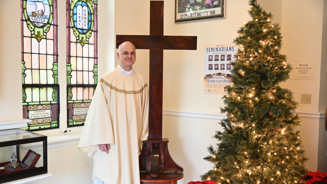 Father Seamus Griesbach standing next to a cross. A Christmas tree is in the corner.